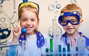 boy and girl standing behind table fll of test tubes.  The girl is holding her finger in the air to show the blue paint her h